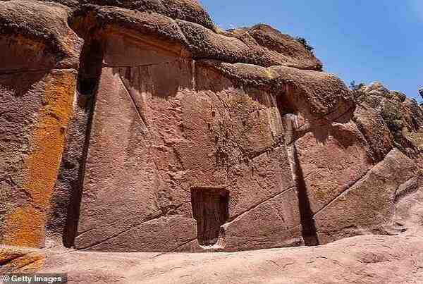 Vedere largă a formațiunii stâncoase cunoscute sub numele de Aramu Muru sau Poarta Zeilor, situată în zona Hayu Marca din Peru, lângă Lacul Titicaca. Imaginea arată o fațadă sculptată cu precizie într-un perete de gresie roșiatică, prezentând o nișă mare în formă de T și o mică adâncitură centrală dreptunghiulară, asemănătoare unei uși.
