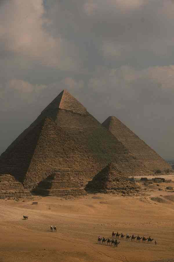 Vedere panoramică a Platoului Giza sub un cer înnorat. Piramidele lui Keops și Kefren domină peisajul deșertic, în timp ce o caravană de cămile și turiști, vizibili în partea de jos, subliniază scara monumentală a structurilor antice în raport cu omul.