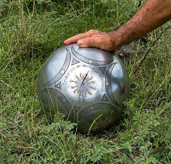 O mână atinge o sferă metalică argintie, cu modele geometrice gravate și un simbol central asemănător unui circuit, care stă în iarbă.